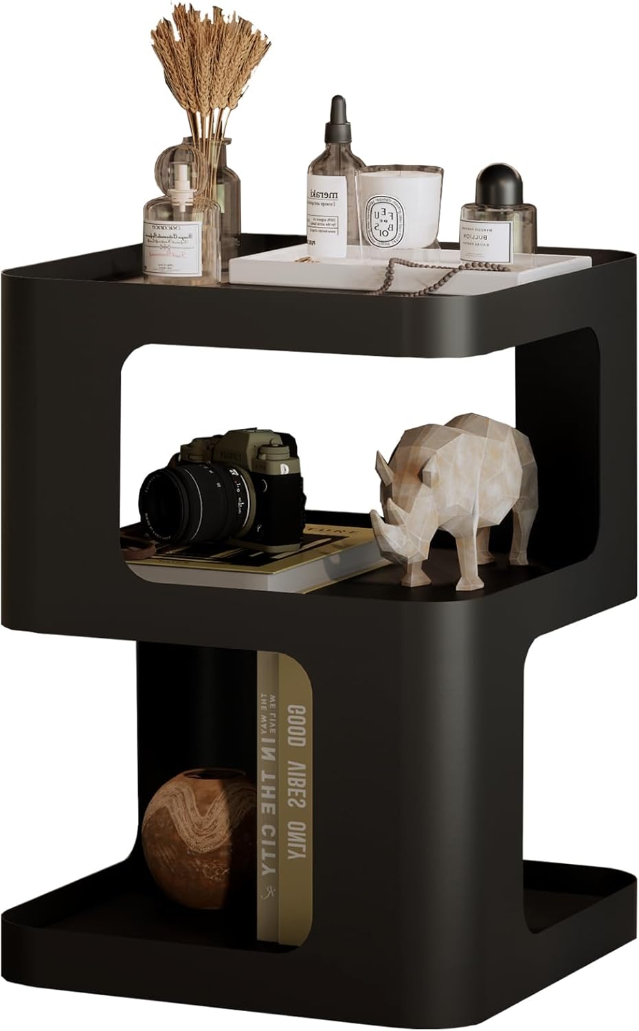 A modern black two-tier side table with open cubby shelves, styled with home decor items including a camera, books, a small rhinoceros figurine, and bottles, shown against a plain white background.