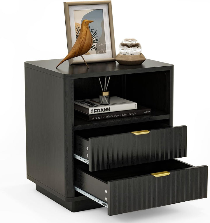 A modern black nightstand with two ribbed drawers featuring gold pull handles, shown with one drawer open. The nightstand is styled with a framed bird print, a small decorative object, and books on its top shelf, set against a plain white background.
