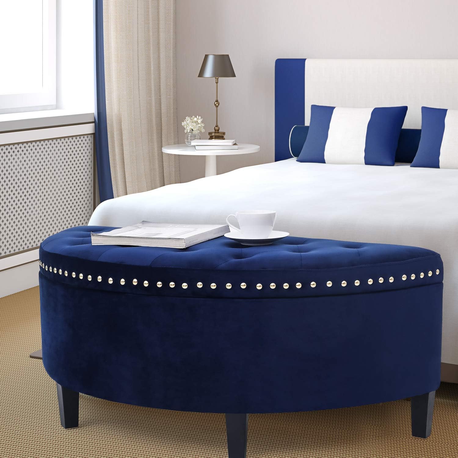 Navy blue ottoman with decorative studs in front of a bed with blue and white pillows.