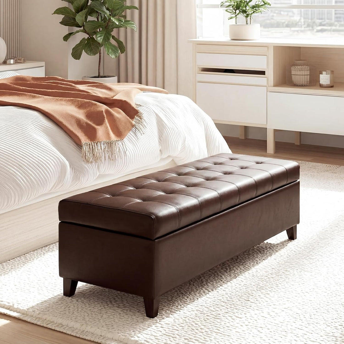 Brown leather ottoman in a bedroom setting with a bed and dresser in the background.