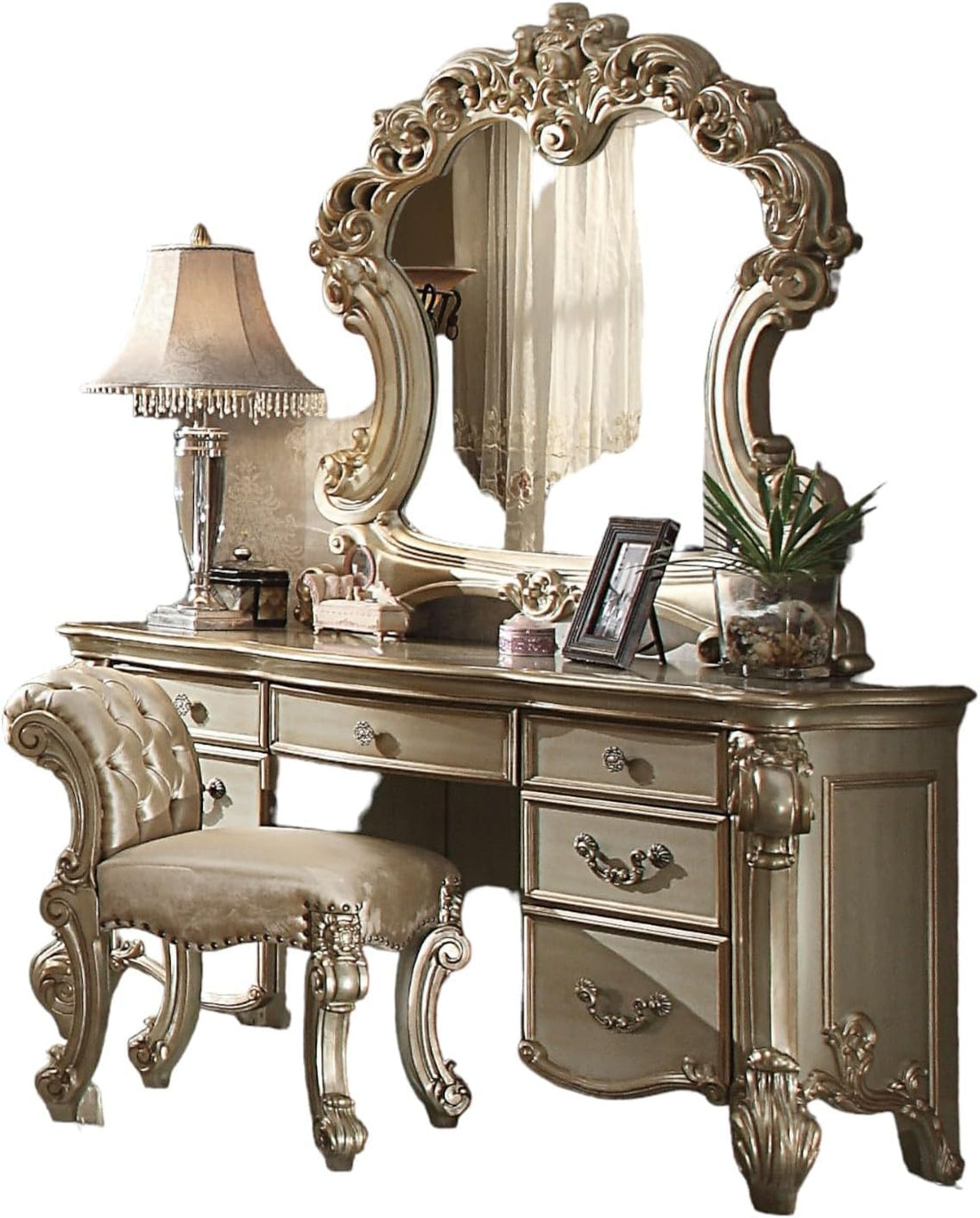 An ornate vanity table set with a large decorative mirror, multiple drawers, a tufted matching stool, and styled accessories including a lamp, framed photo, and small plant, shown on a plain white background.