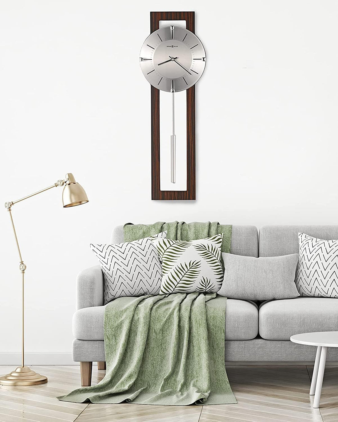 A modern wall clock with a silver circular face and dark wood frame hangs above a light gray sofa in a styled living room setting with a green throw blanket, patterned pillows, and a brass floor lamp.