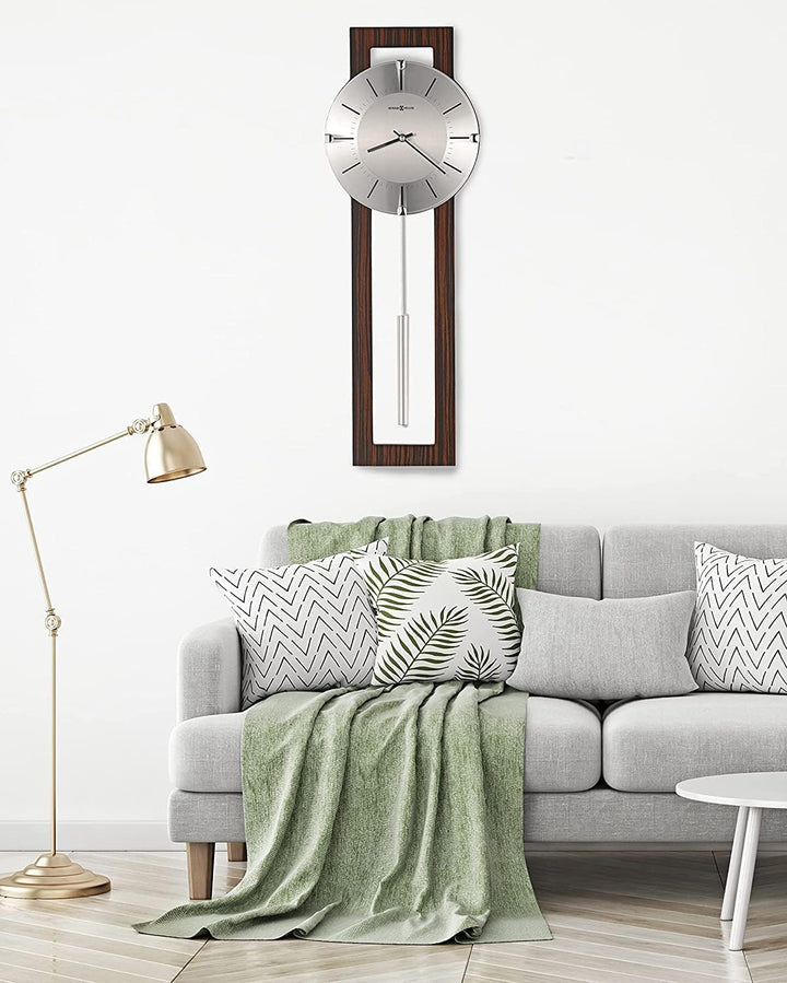 A modern wall clock with a silver circular face and dark wood frame hangs above a light gray sofa in a styled living room setting with a green throw blanket, patterned pillows, and a brass floor lamp.