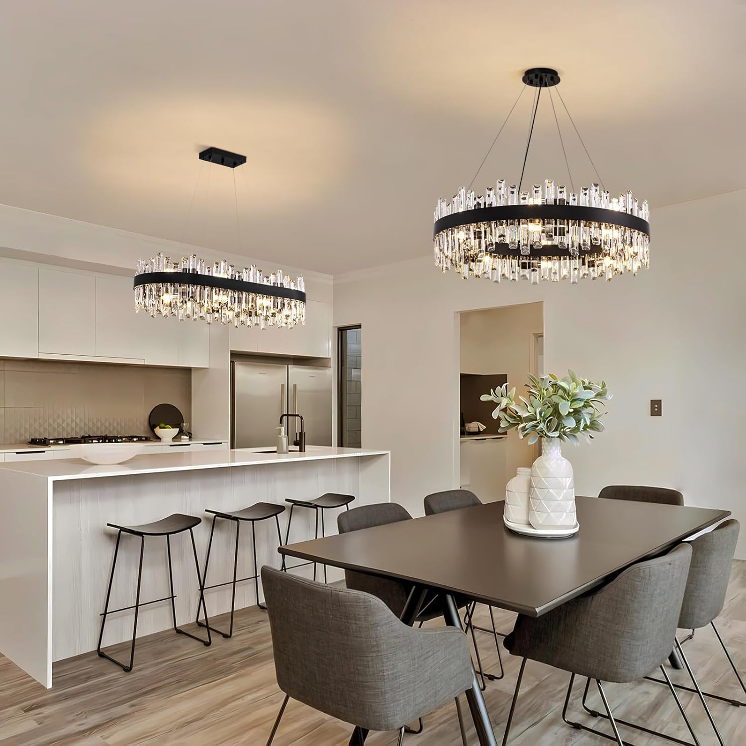 A modern dining room and kitchen interior featuring two large circular chandeliers with clear hanging crystals and black rings, a dark wood dining table, gray upholstered chairs, and a white island bar in the background.