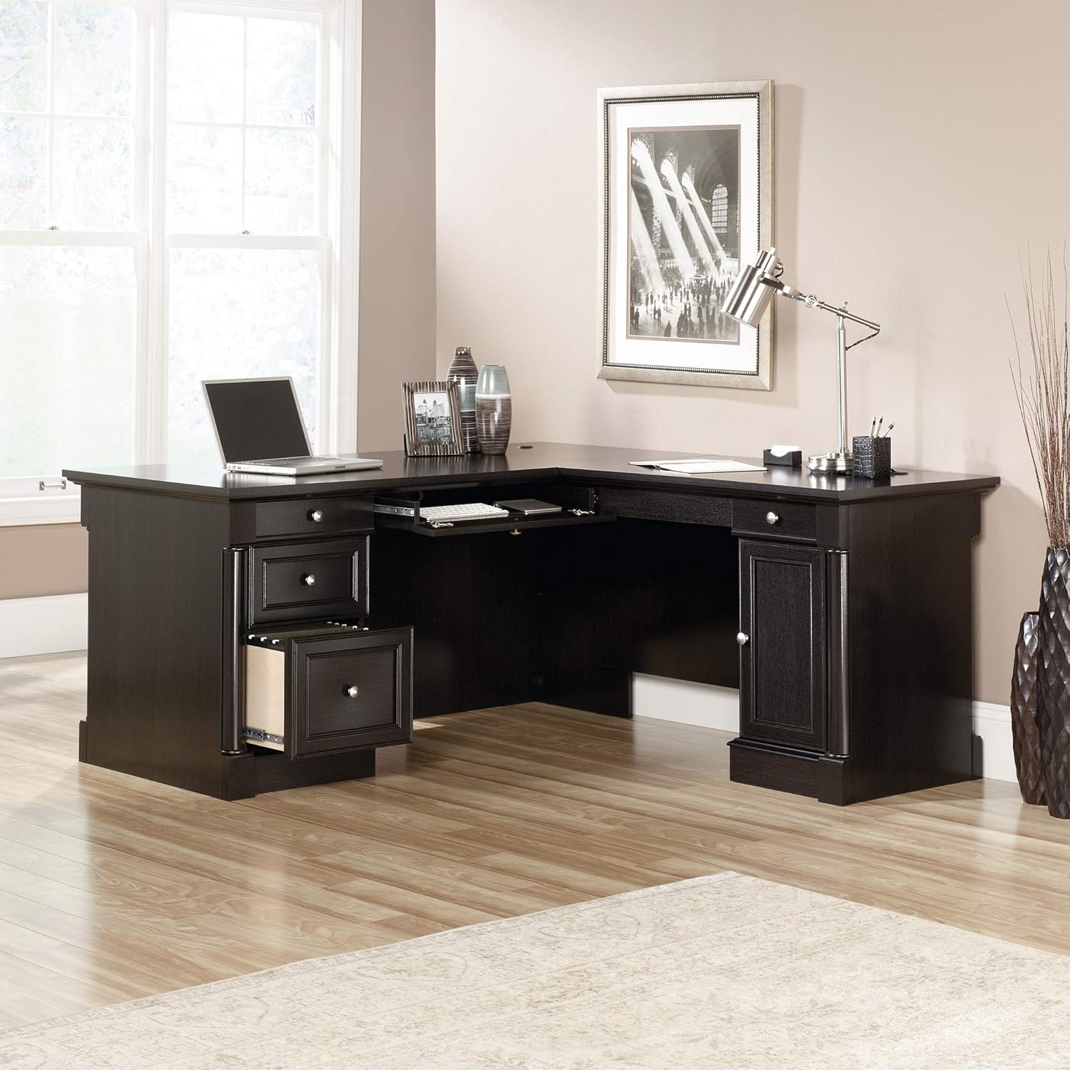 Dark wood L-shaped desk in a home office setting with a laptop, lamp, and decor items.