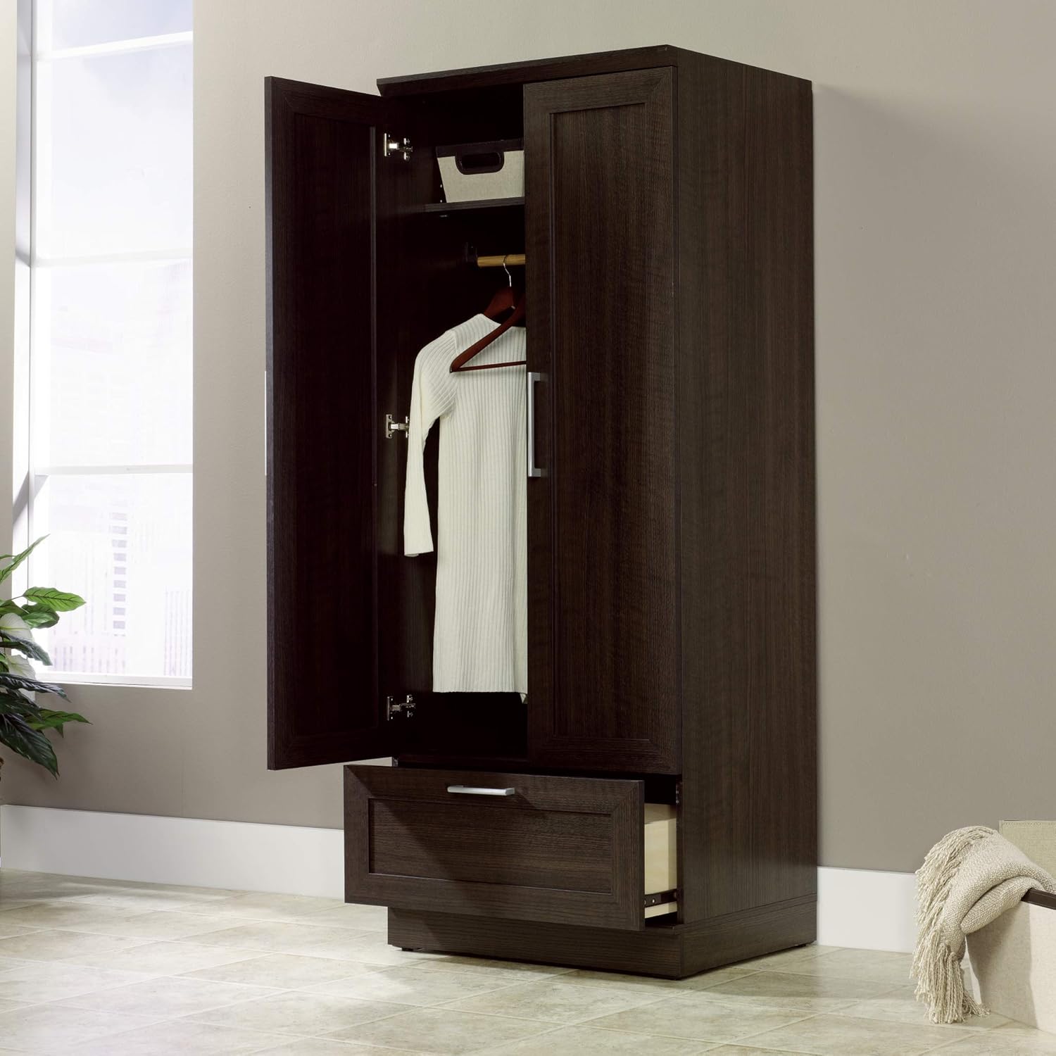 Dark wooden wardrobe with open doors showing clothes in a room with a window and plant.