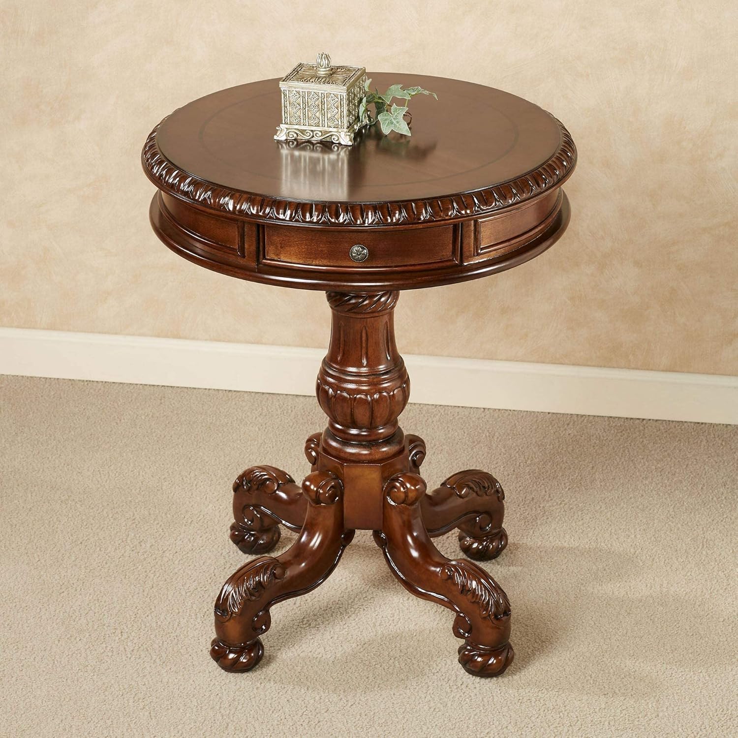A round wooden pedestal side table with carved details, a single drawer, and ornate claw-style legs, shown in a warm brown finish against a beige wall and carpet.