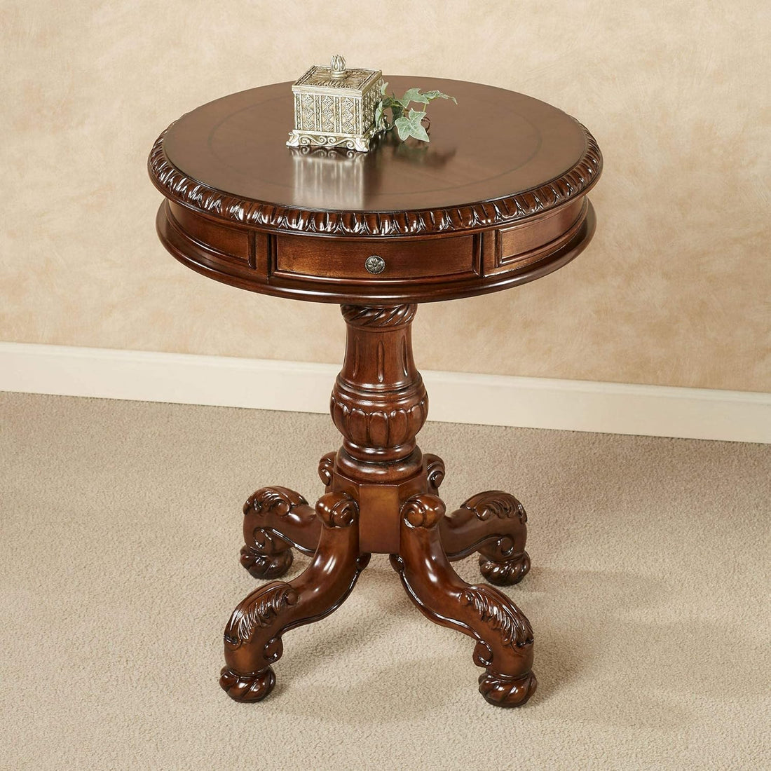 A round wooden pedestal side table with carved details, a single drawer, and ornate claw-style legs, shown in a warm brown finish against a beige wall and carpet.