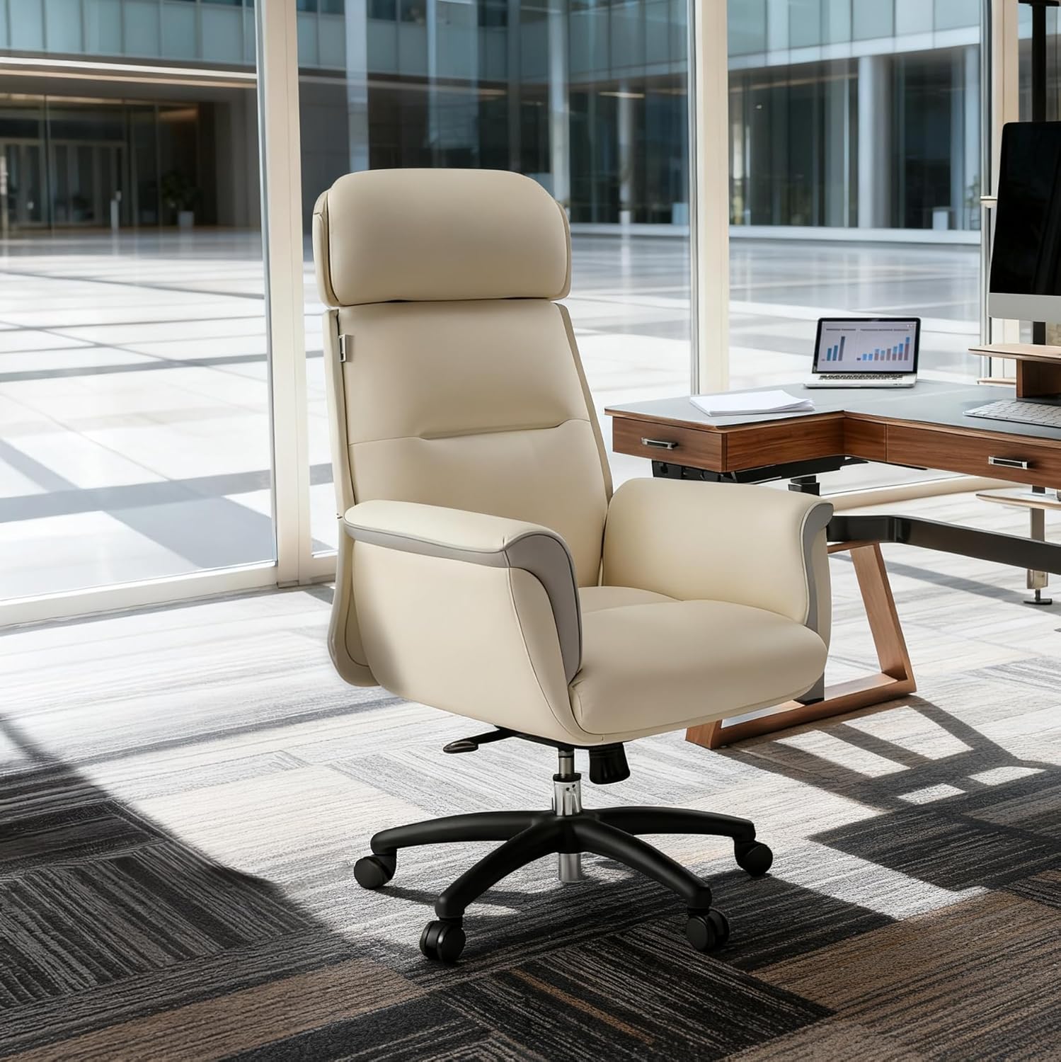 A cream-colored high-back office chair with a black five-star wheeled base, positioned in a modern office setting with a desk and large windows in the background.
