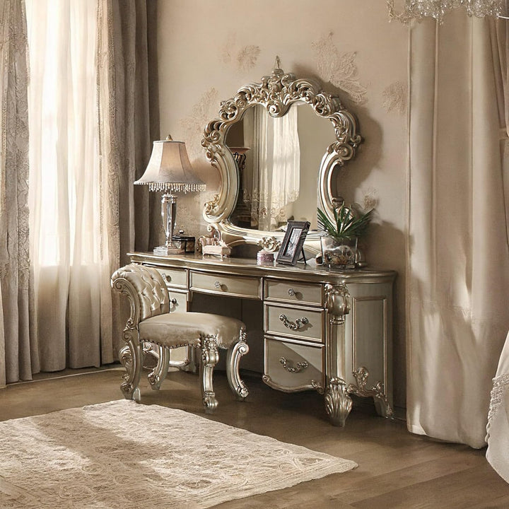 An ornate silver vanity table with a large decorative mirror, matching upholstered stool, and a table lamp in a luxurious beige-toned bedroom setting.