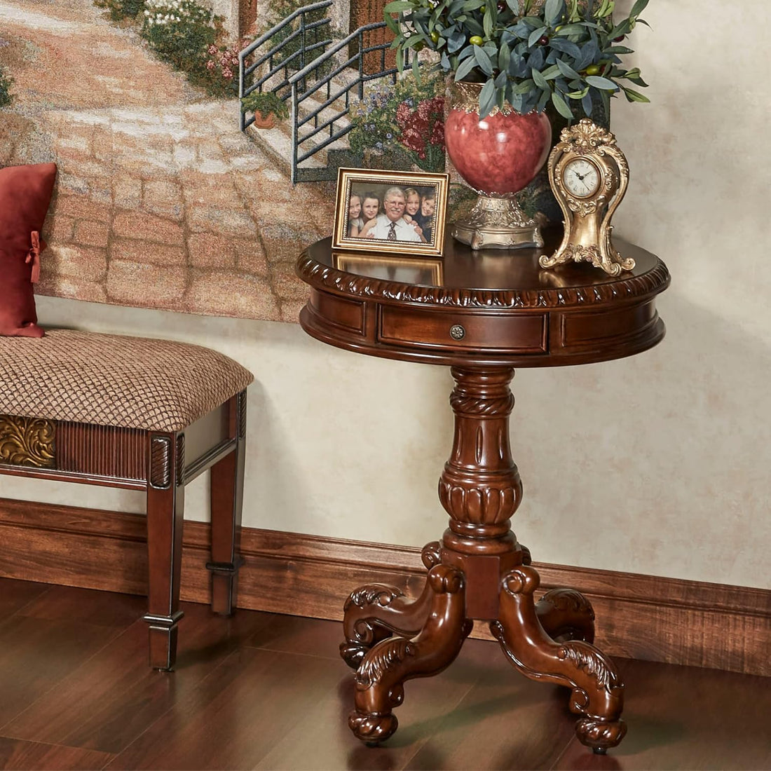An ornate round wooden end table with carved details and three legs, styled in a home interior next to a bench, holding decorative items including a red vase, a small framed photo, and a gold clock.