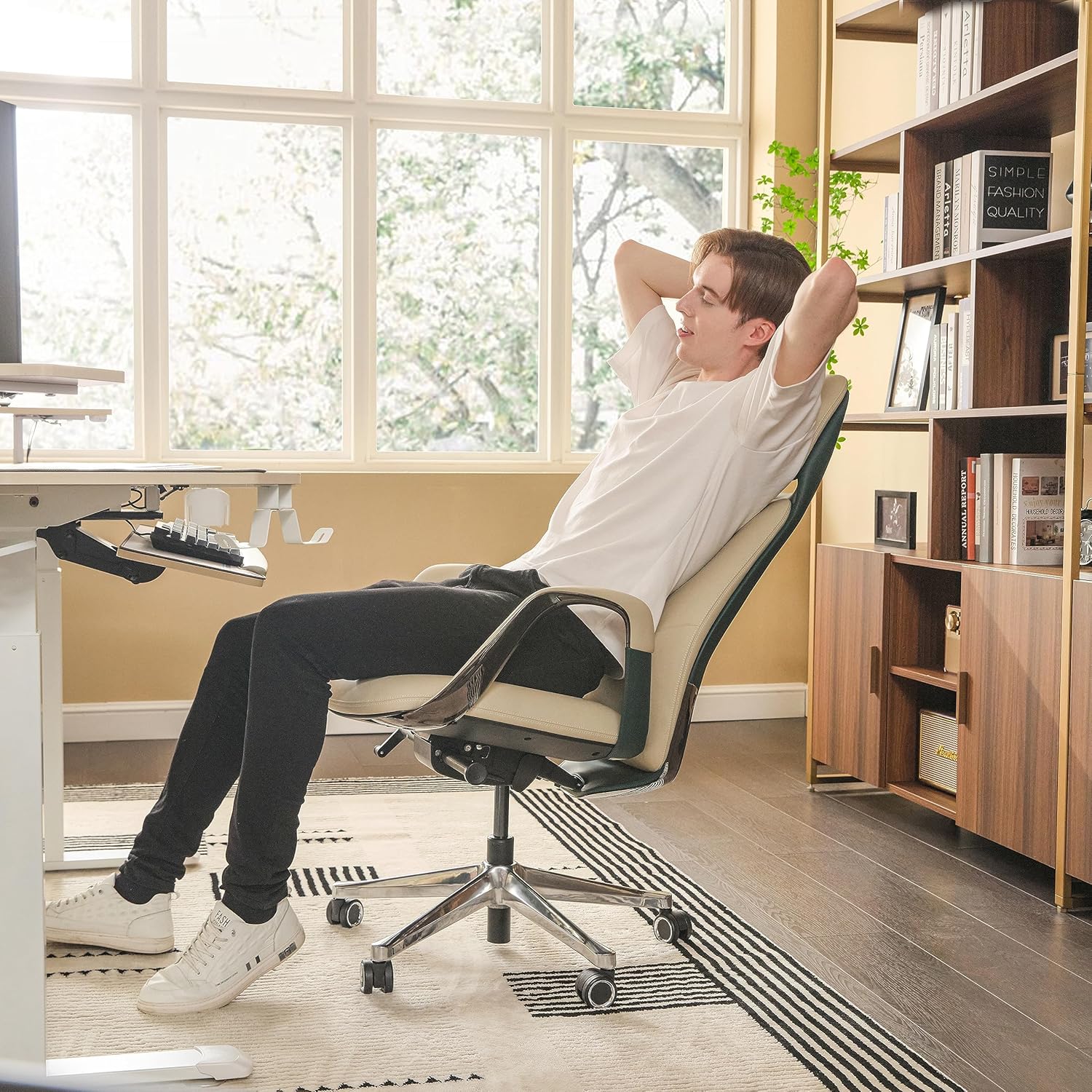 Person relaxing on a chair in a home office setting with large windows and bookshelves.