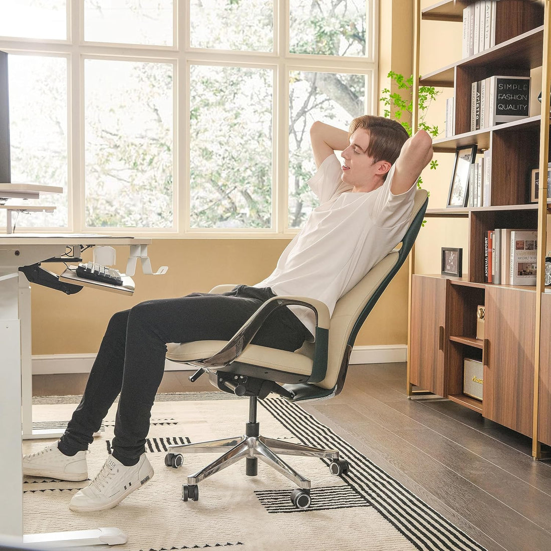 Person relaxing on a chair in a home office setting with large windows and bookshelves.