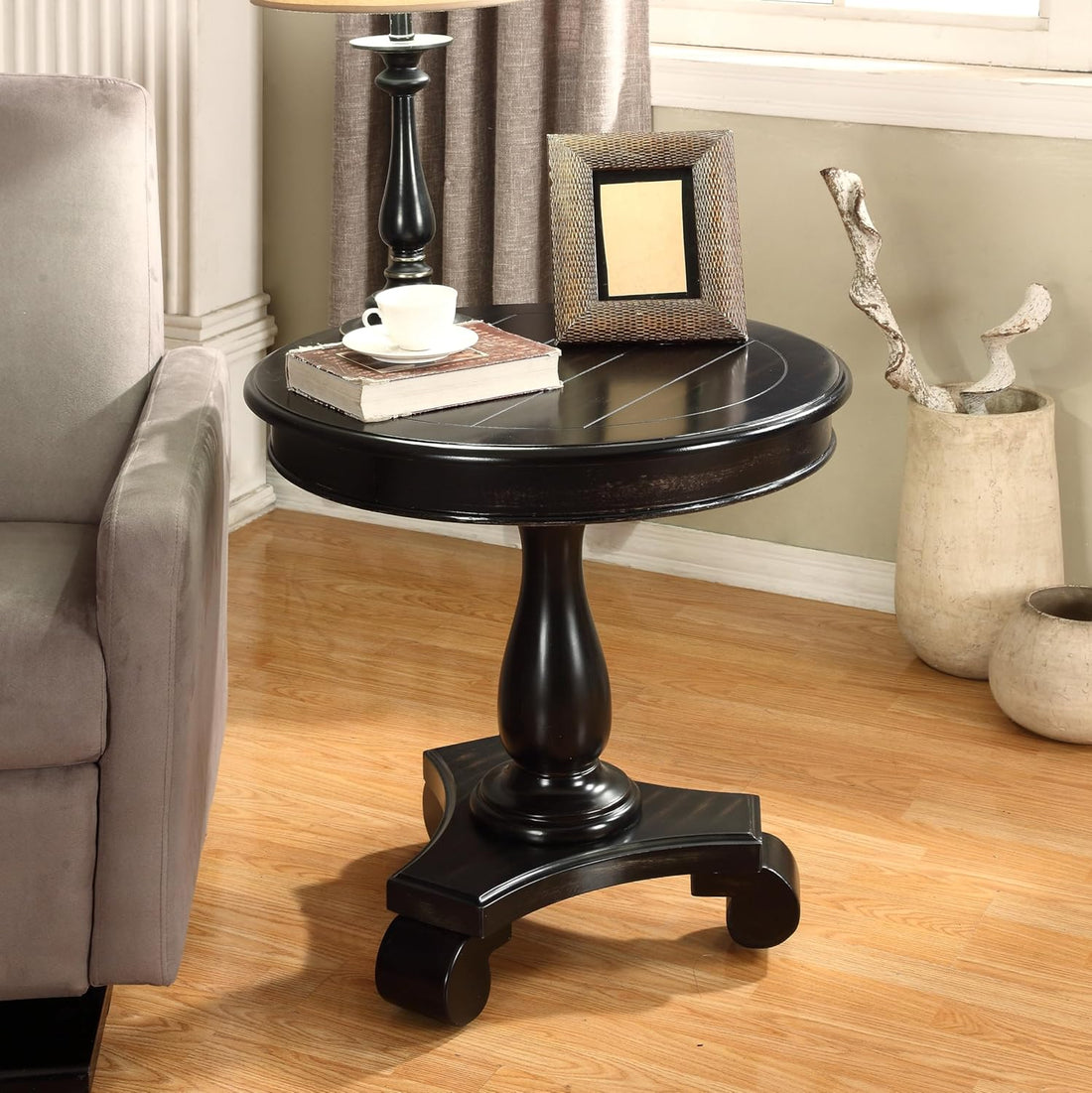 A round black wooden end table with a turned pedestal base and casters, styled in a living room next to a gray armchair with books, a picture frame, and a lamp.
