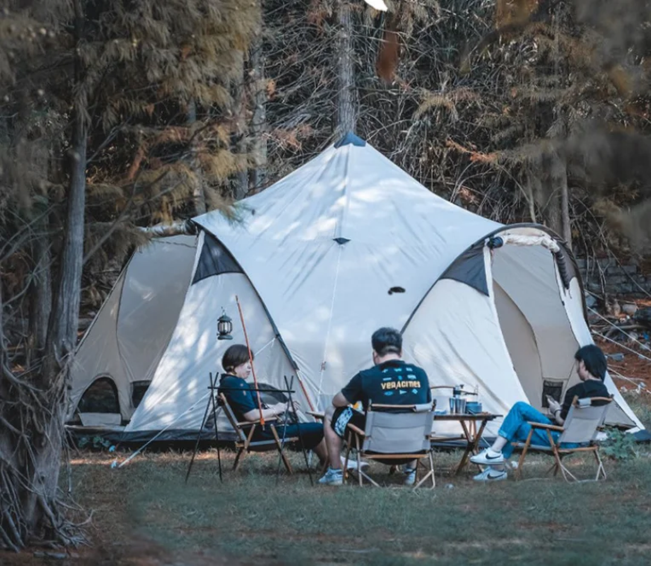 Three people sitting around a table outside a large tent in a forest setting.
