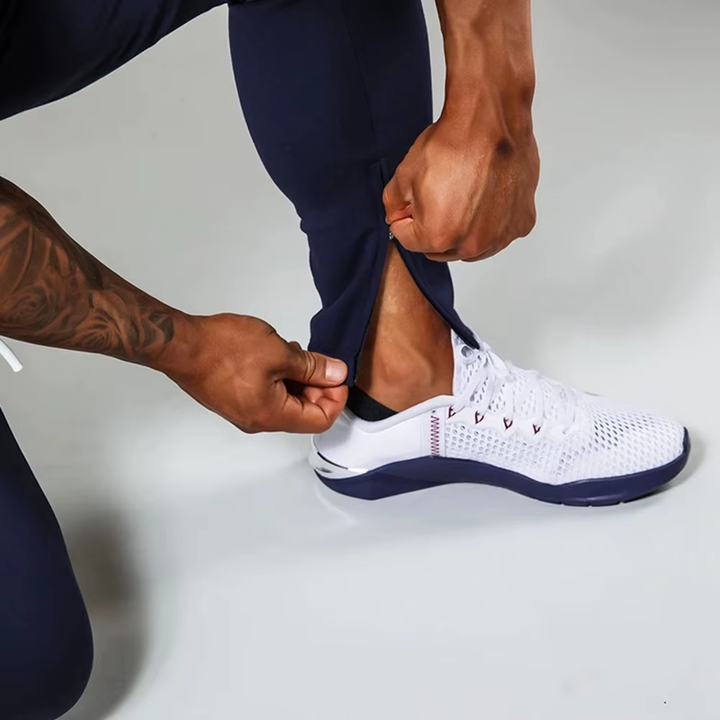 A close-up of a person pulling up navy athletic pants to reveal a white athletic shoe with navy trim, shown against a light gray studio background.