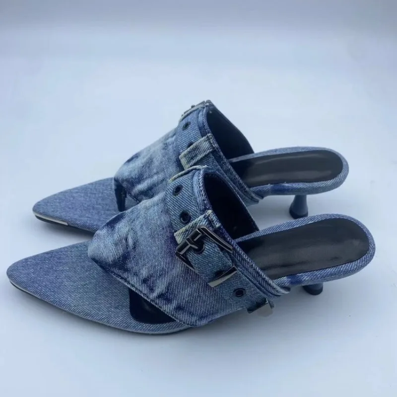 A pair of pointed-toe mule sandals made from blue denim, featuring a wide strap with a silver buckle and a short dark heel, shown on a light gray background.