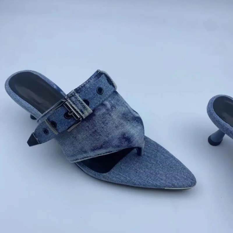 A single pointed-toe mule sandal with a denim-like blue fabric upper and a small metallic buckle detail, photographed on a plain light background. A second shoe is partially visible at the right edge.