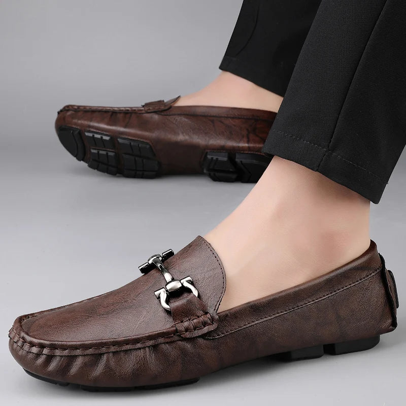 Brown leather loafers with a metallic buckle worn by a person on a gray background