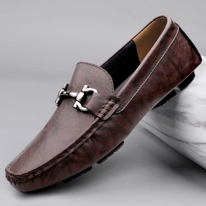 Brown leather loafer with a silver buckle on a gray background