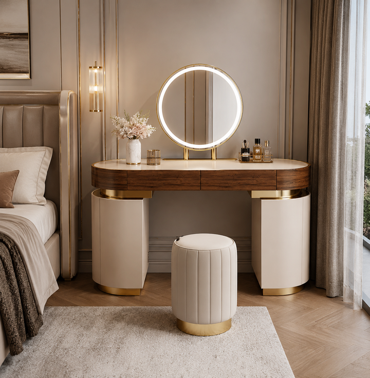 A modern vanity table set in a bedroom, featuring a round backlit LED mirror, a dark wood top with cream marble-look front panels, and gold metal accents. A matching cream and gold velvet stool sits in front.