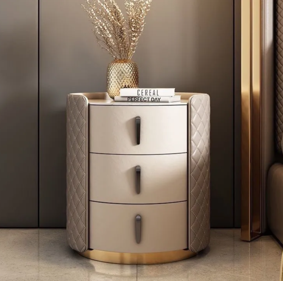 A modern three-drawer nightstand with champagne-colored drawers, quilted taupe side panels, and a gold base, styled with a vase of dried grass and books in a luxury bedroom setting.