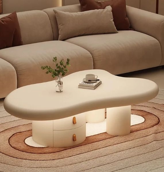 A modern cloud-shaped coffee table with integrated drawers and warm under-glow lighting, styled in a cozy living room setting with a beige sofa and patterned rug.