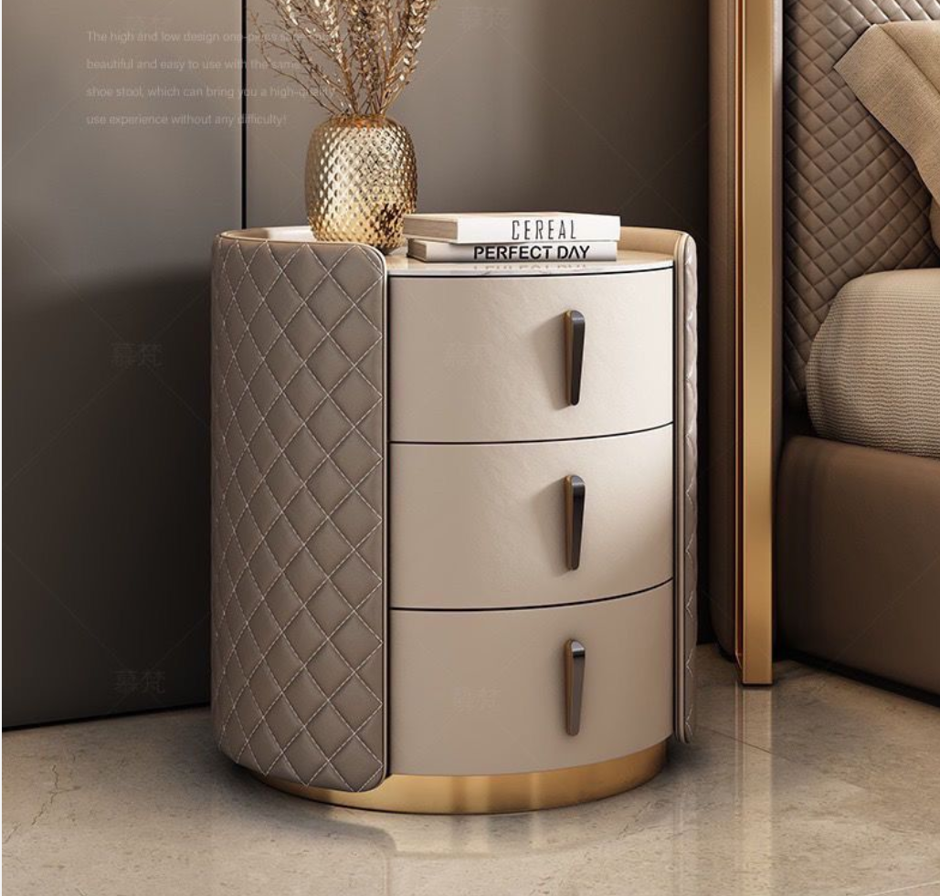 A modern three-drawer nightstand with a cream-colored front, quilted taupe side panels, and gold metal handles and base trim, styled in a bedroom next to a bed.