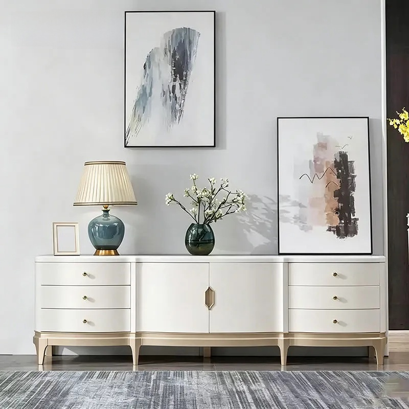 A modern white sideboard console table with gold-toned legs and drawer pulls, styled in a minimalist room with a blue-green ceramic table lamp, a vase of white blossoms, and two framed abstract prints on a light gray wall.