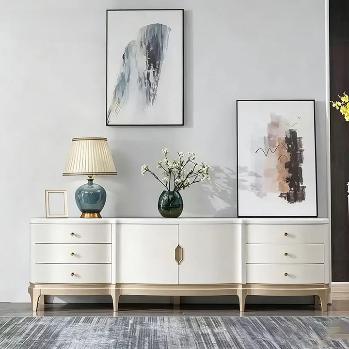 A modern white sideboard console table with gold-toned legs and drawer pulls, styled in a minimalist room with a blue-green ceramic table lamp, a vase of white blossoms, and two framed abstract prints on a light gray wall.