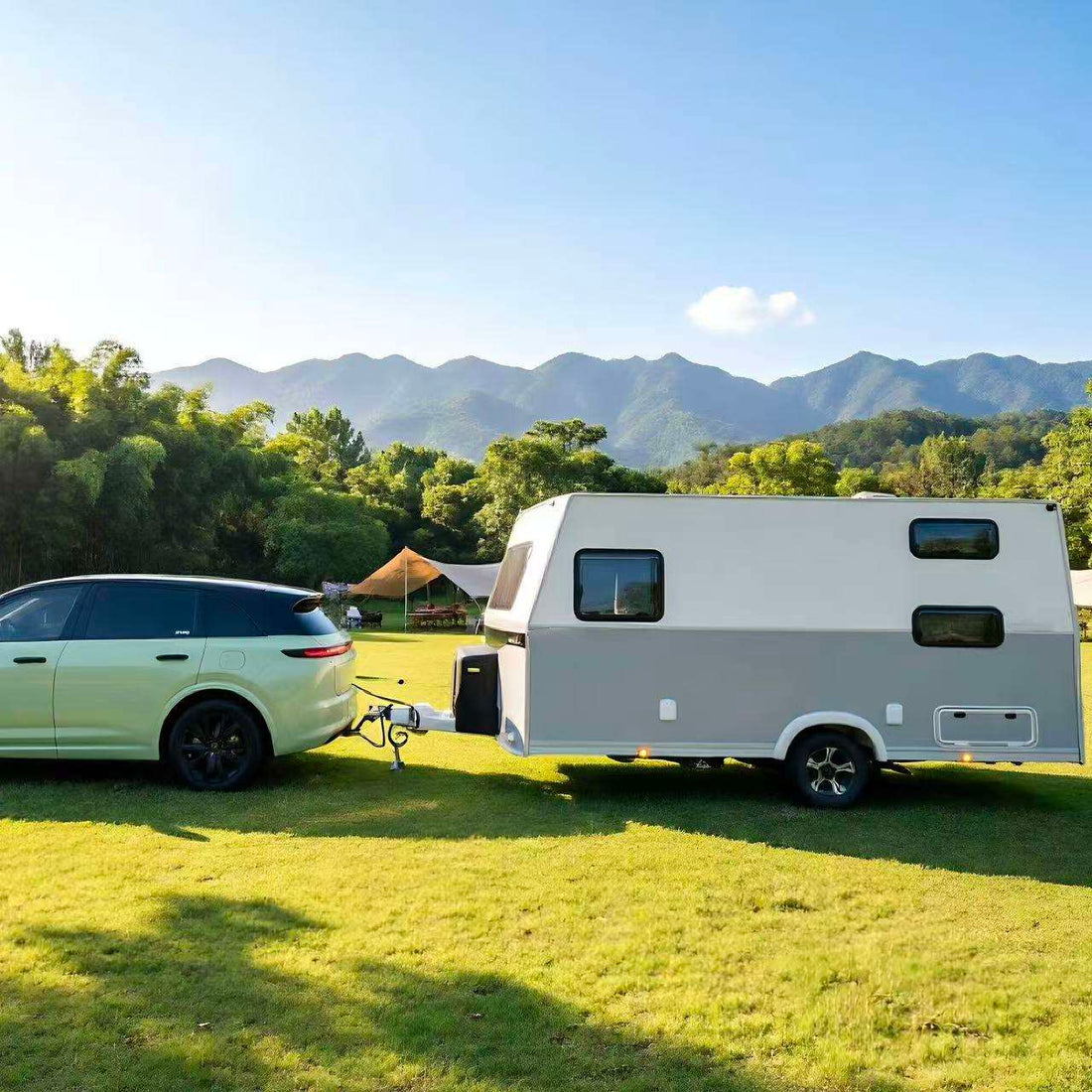 A white and gray travel trailer parked on a grassy field, hitched to a light green SUV, with mountains and trees in the background.