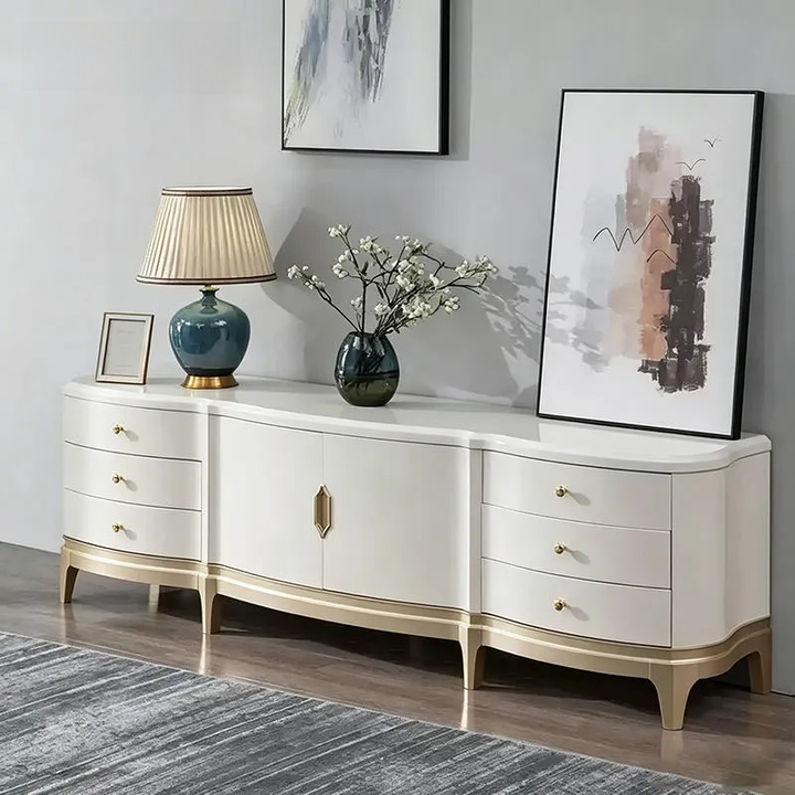 A modern curved white sideboard or credenza with gold-toned legs and drawer pulls, styled in a living room setting with a blue table lamp, a vase of white blossoms, and framed wall art.