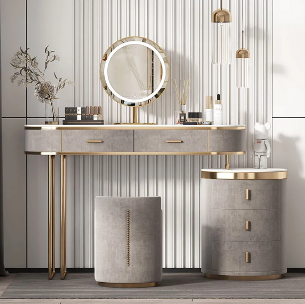 A modern vanity table set with a gray upholstered console table, matching stool, and side cabinet with gold-tone hardware, shown against a vertically paneled wall with decorative accessories.