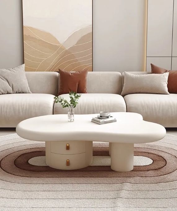 A modern living room scene featuring a cream-colored, cloud-shaped coffee table with two drawers and gold handles, set on a striped area rug in front of a beige sofa with brown accent pillows.
