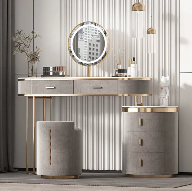 A modern vanity table set with a round backlit mirror, a gray upholstered stool, and a matching gray three-drawer side cabinet with gold trim, set against a vertical slatted gray wall.