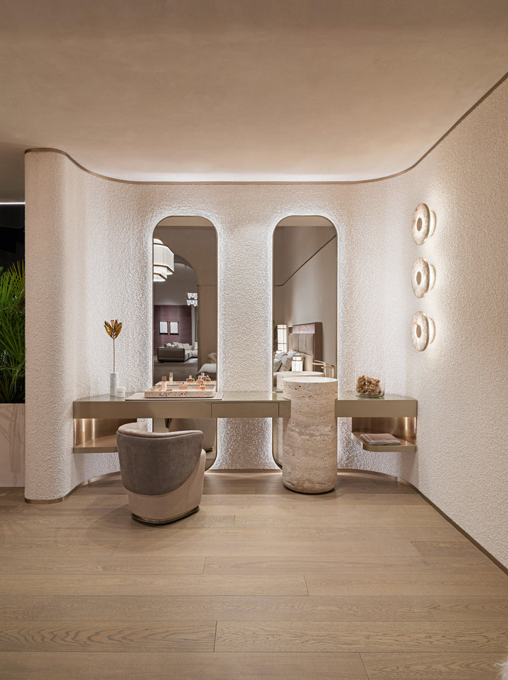 A luxury modern vanity area with a long floating desk, two arched mirrors, a textured gray and beige accent chair, and warm ambient lighting on a light wood floor.