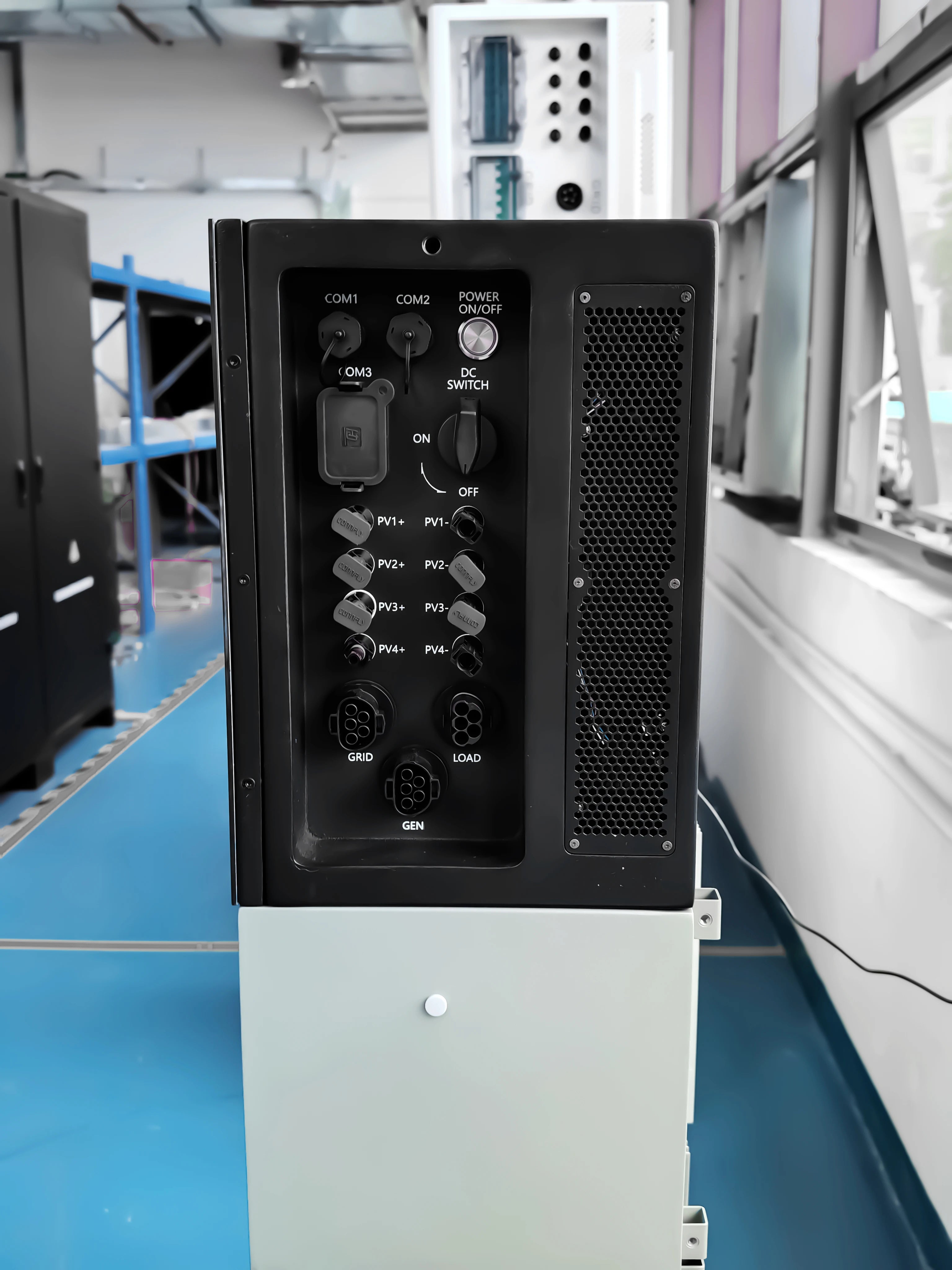 A black industrial power supply or generator control panel mounted on a light gray cabinet base, photographed in a workshop or server room with blue flooring and other equipment visible in the background.