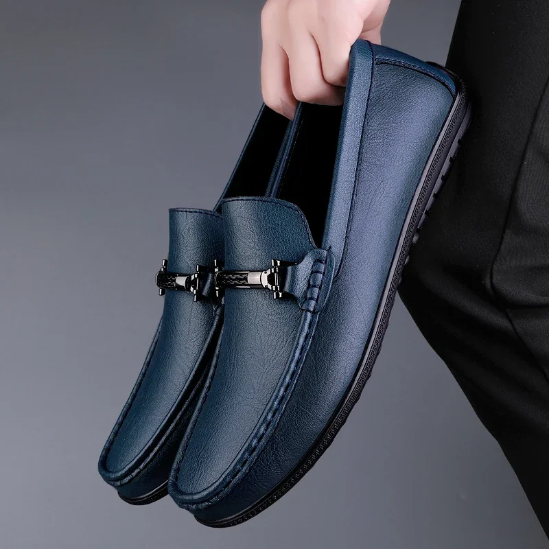Blue loafers with a gold buckle held by a hand against a gray background
