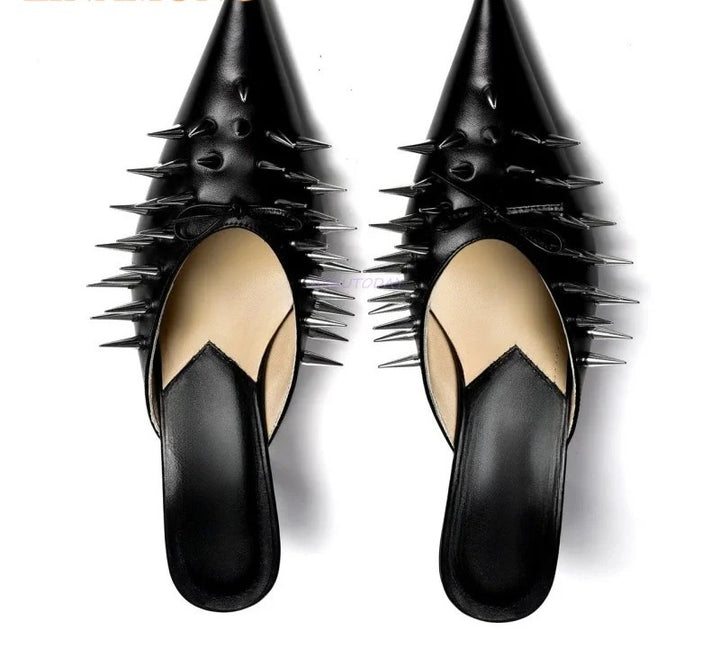 A pair of black pointed-toe mule flats with prominent metallic spikes along the sides, shown from a top-down view on a white background.