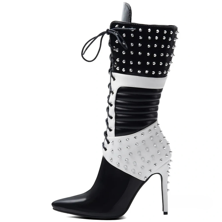 A knee-high studded boot with a pointed toe and high stiletto heel, featuring a black and white color-blocked design with silver-tone studs and side laces, shown against a plain white background.