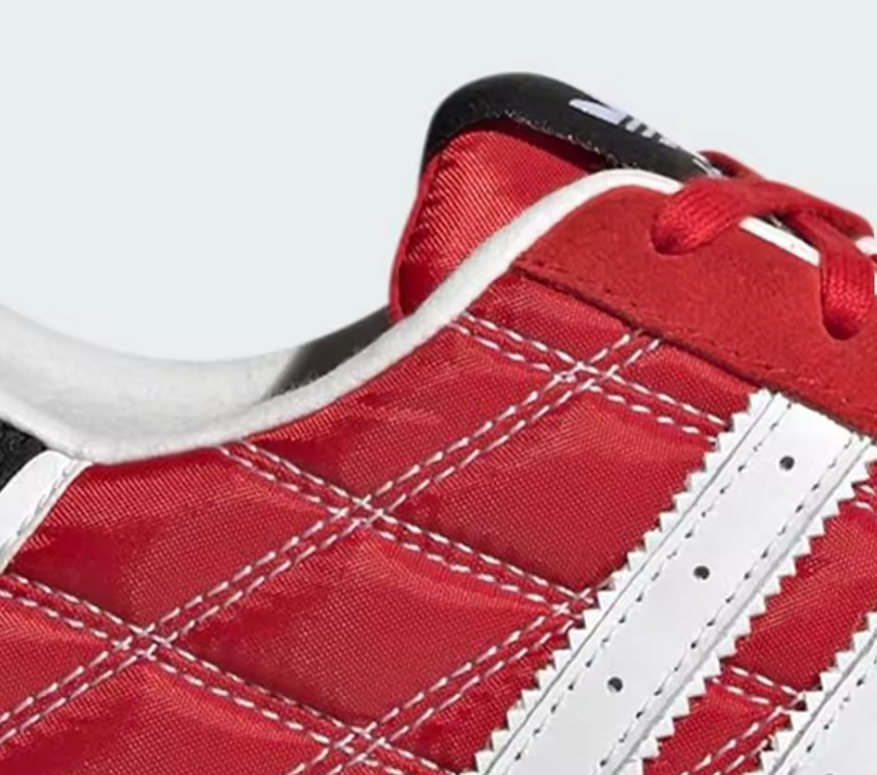 Close-up of a red Adidas sneaker showing the side panel with white contrast stitching, white stripes, red laces, and a black heel tab on a light gray background.